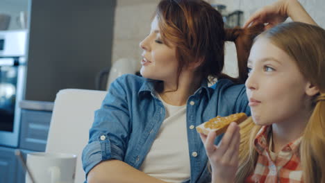 Portrait-shot-of-the-pretty-woman-and-her-blonde-teen-daughter-sitting,-talking-and-resting-while-a-girl-eating-bread-with-peanut-butter.-Close-up.-Indoors
